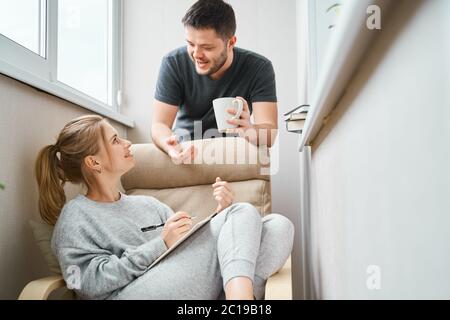 Donna sorridente seduta su poltrona beige e uomo brunetto in piedi accanto a lei sul balcone Foto Stock