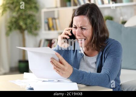 Triste donna adulta che piange chiamando su smartphone con lettera seduta sul pavimento a casa Foto Stock