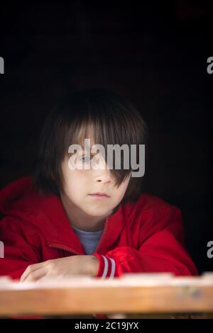 Ragazzo di preda in felpa rossa, nascosto dietro una porta di legno, che guarda spaventato e triste Foto Stock