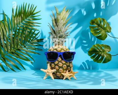Fresco ananas hipster in occhiali da sole tra stelle del mare, palme e foglie di monstera su sfondo blu. Forti ombre tropicali. Vacanza hawaiana in spiaggia Foto Stock