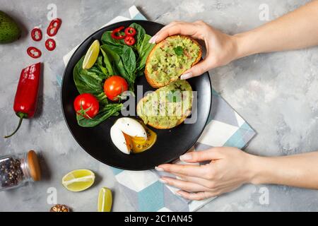 Mano femminile che tiene un sano toast con avocado messicano salsa guacamole, uova bollite, pomodori ciliegini e insalata su fondo di cemento. Vista dall'alto. Brea Foto Stock