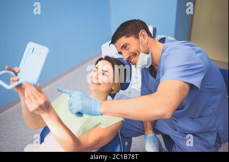Il dentista mostra al paziente i risultati del trattamento con uno specchio, esaminando i denti con l'apparecchiatura dentale in studio dentistico. Giovane bel medico maschile Foto Stock