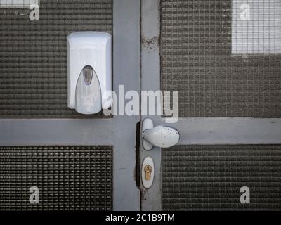 Dispenser di igienizzante per mani montato sulla parete della porta d'ingresso con serratura e manopola, in luogo pubblico, un edificio residenziale, che distribuisce idroalcool Foto Stock