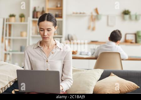 Ritratto dai toni caldi di una giovane donna elegante che usa il computer portatile a casa con suo figlio che studia in background, spazio di copia Foto Stock