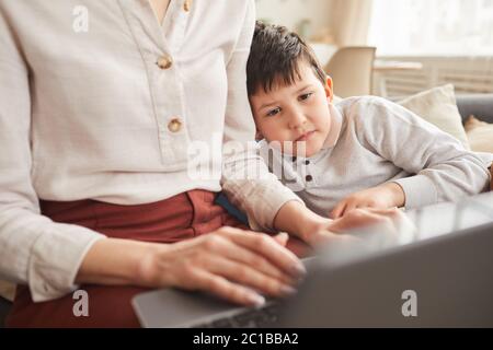Primo piano ritratto di un ragazzo carino che guarda lo schermo del computer portatile mentre studia a casa con madre che lo aiuta in interni accoglienti, spazio di copia
