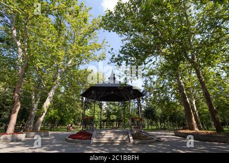 Chiosco in un parco cittadino in estate, circondato da giardini e alberi alti, con il suo tipico palco rotondo e rifugio in metallo. Un chiosco o un chiosco musicale, Foto Stock