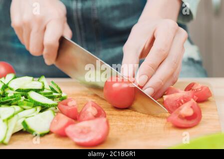 Primo piano le mani femminili delicate tagliano un coltello grande con pomodori su un quarto su una tavola di legno a casa. Cucina casala. Mangiare sano Foto Stock