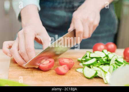 Primo piano le mani femminili delicate tagliano un coltello grande con pomodori su un quarto su una tavola di legno a casa. Cucina casala. Mangiare sano Foto Stock