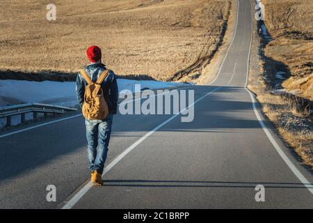 Un hipster con un vecchio zaino vintage indossando occhiali da sole con un cappello rosso e una giacca jeans e jeans a piedi vanno Foto Stock