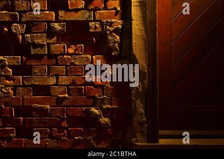 Vecchio grugnoso e danneggiato muro di mattoni rossi di notte accanto ad una porta di legno con luce di una lanterna che splende su di essa Foto Stock