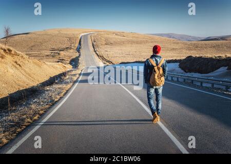 Un hipster con un vecchio zaino vintage indossando occhiali da sole con un cappello rosso e una giacca jeans e jeans a piedi vanno Foto Stock