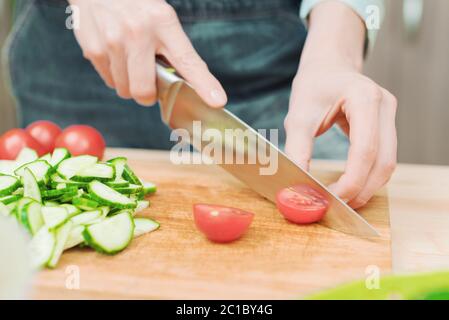 Primo piano le mani femminili delicate tagliano un coltello grande con pomodori su un quarto su una tavola di legno a casa. Cucina casala. Mangiare sano Foto Stock