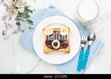 Food Art Brindisi con orso per bambini con crema di nocciole al cioccolato, frutta e bicchiere di latte. Vista dall'alto su un asciugamano blu Foto Stock