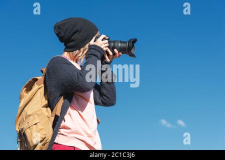 Ritratto di una ragazza elegante hippy in un cappello e con uno zaino che scatta le foto di lei su una fotocamera DSLR all'aperto contro un blu Foto Stock