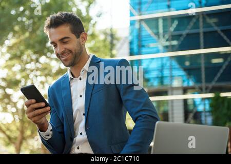 Uomo d'affari sorridente seduto fuori a leggere un testo sul suo cellulare Foto Stock