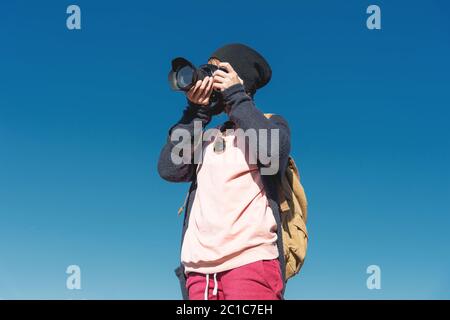 Ritratto di una ragazza elegante hippy in un cappello e con uno zaino che scatta le foto di lei su una fotocamera DSLR all'aperto contro un blu Foto Stock