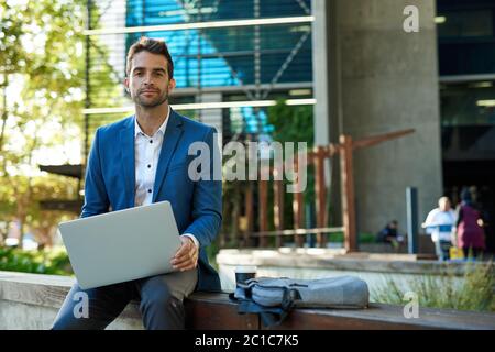 Uomo d'affari sicuro seduto fuori dall'edificio dell'ufficio utilizzando un computer portatile Foto Stock