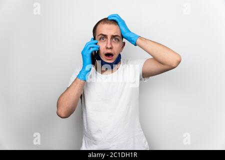 Uomo che parla al telefono ricevendo notizie molto cattive e tristi, espressione molto preoccupata, indossando guanti di protezione blu e maschera viso sul mento. Bianco shi Foto Stock