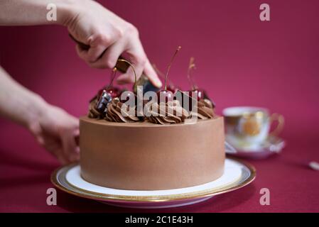 Torta al cioccolato con ciliegia e una tazza di caffè. Le mani femminili tagliano una torta con ciliegia. Foto Stock