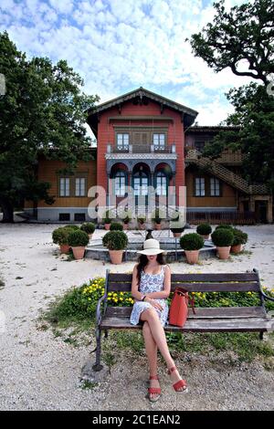 Giovane donna seduta su una panchina al parco di Villa Revoltella con sullo sfondo lo chalet in stile liberty Foto Stock