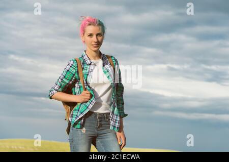 ritratto di una giovane donna sorridente attraente in abiti da camicia plaid con piccolo zaino in giornata di sole sul cielo nuvoloso e campo Foto Stock