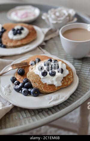 Colazione sana su un tavolo da caffè in metallo. Frittelle di farinata d'avena ricoperte di yogurt e mirtilli e una tazza di caffè sfocata in lontananza. Foto Stock