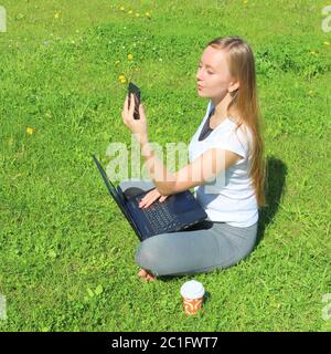 Una bella giovane ragazza bianca in una t-shirt bianca e con i capelli lunghi seduti su erba verde, sul prato e lavorando dietro un portatile nero e guarda il display del telefono. Foto Stock