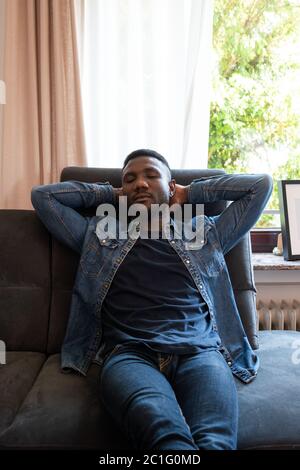 Giovane uomo nero che pende sul divano del soggiorno con le mani dietro la testa. Vista frontale. Lunghezza di tre quarti. Foto Stock