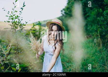 Bella ragazza in una tavola e cappello raccoglie i fiori. La ragazza raccoglie un bouquet di fiori secchi. Bruna ragazza tra l'erba verde alta Foto Stock