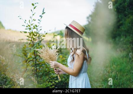 Bella ragazza in una tavola e cappello raccoglie i fiori. La ragazza raccoglie un bouquet di fiori secchi. Bruna ragazza tra l'erba verde alta Foto Stock