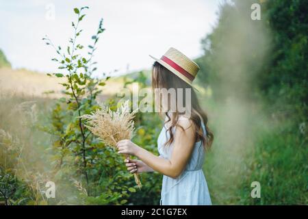 Bella ragazza in una tavola e cappello raccoglie i fiori. La ragazza raccoglie un bouquet di fiori secchi. Bruna ragazza tra l'erba verde alta Foto Stock