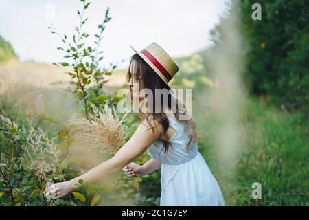 Bella ragazza in una tavola e cappello raccoglie i fiori. La ragazza raccoglie un bouquet di fiori secchi. Bruna ragazza tra l'erba verde alta Foto Stock