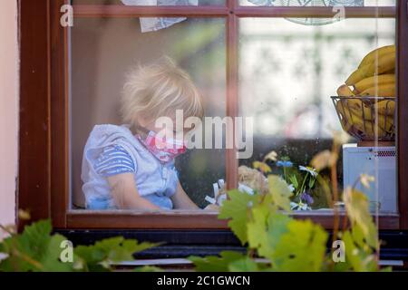 Dolce bambino, indossando maschera medica, orsacchiotto, anche con maschera, guardando tristemente fuori dalla finestra, durante l'isolamento pandemico del coronavirus Foto Stock