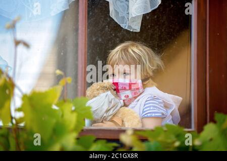 Dolce bambino, indossando maschera medica, orsacchiotto, anche con maschera, guardando tristemente fuori dalla finestra, durante l'isolamento pandemico del coronavirus Foto Stock