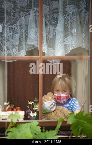 Dolce bambino, indossando maschera medica, orsacchiotto, anche con maschera, guardando tristemente fuori dalla finestra, durante l'isolamento pandemico del coronavirus Foto Stock