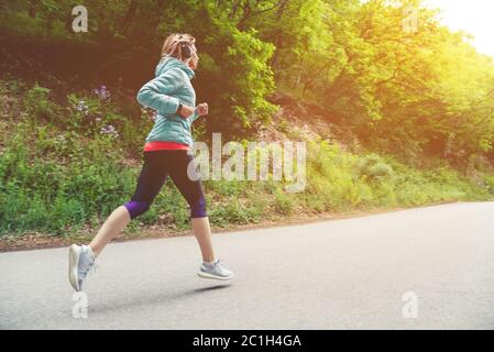 Una giovane bionda che corre pratica all'aperto in un parco cittadino di montagna nella foresta. Raggi caldi attraverso i rami di tre Foto Stock