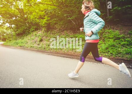 Giovane donna bionda fitness in cuffia che corre al mattino percorso forestale caucasico in luce solare Foto Stock