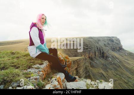 Una ragazza-viaggiatore con capelli multicolore si siede sul bordo di una scogliera e guarda all'orizzonte su uno sfondo di un altopiano roccioso Foto Stock