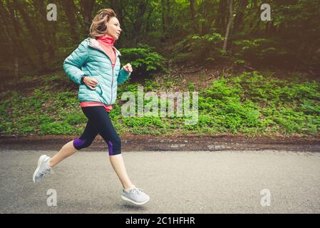 Una giovane bionda che corre pratica all'aperto in un parco cittadino di montagna nella foresta. Raggi caldi attraverso i rami di tre Foto Stock