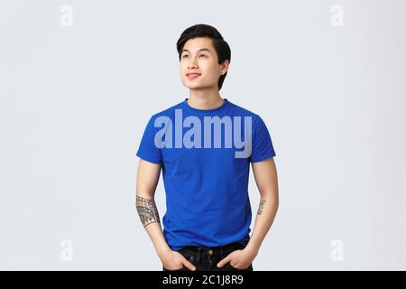 Sognante e premuroso ragazzo di queer asiatico in t-shirt blu, guardando qualcosa in avanti, guardando in alto a sinistra con un viso allegro e sorridente Foto Stock