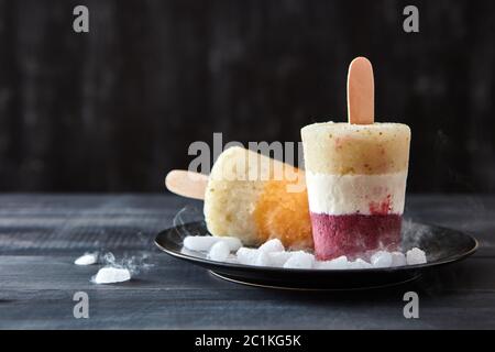 Vari caffè, gelato alla frutta su un bastone sono presentati in un piatto nero con fette di ghiaccio su uno sfondo di legno scuro con sp Foto Stock