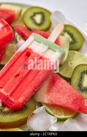 Primo piano frutta surgelati su un bastone sotto forma di un pezzo di anguria in un piatto con cubetti di ghiaccio e pezzi di frutta su Foto Stock