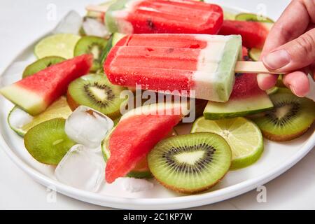 La mano della donna in primo piano tiene il gelato alla bacca in lolly, su uno sfondo bianco un piatto di gelato, pezzi di kiwi, anguria e ic Foto Stock