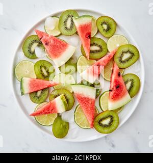 Succose fette di frutta, calce, kiwi, anguria e cubetti di ghiaccio in una piastra su un marmo grigio Sfondo. Lay piatto Foto Stock