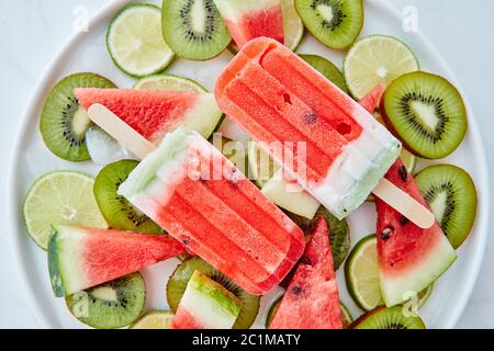Frappè di anguria surgelati su un bastone in una ciotola con pezzi di kiwi, lime, anguria e fette di ghiaccio su un dorso di marmo grigio Foto Stock