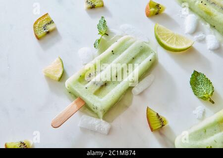 Succo di frutta surgelato su un bastone con un pezzo di kiwi fresco su uno sfondo di marmo grigio con foglie di menta e pezzi di lime, ghiaccio e Foto Stock