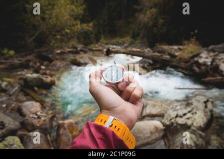 Una bella mano maschile con una cinghia gialla di orologio tiene una bussola magnetica in una foresta di conifere autunno contro un fiume di montagna Foto Stock