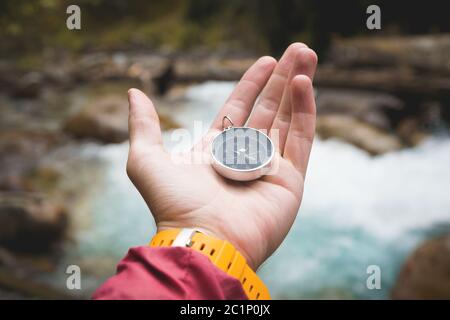 Una bella mano maschile con una cinghia gialla di orologio tiene una bussola magnetica in una foresta di conifere autunno contro un fiume di montagna Foto Stock