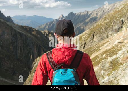 Un uomo bearded in un cappello con uno zaino si trova sulla cima di una roccia e si affaccia in una valle rocciosa alta in montagna. Il concetto Foto Stock