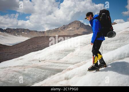 Un alpinista con uno zaino cammina in ramponi camminando lungo un ghiacciaio polveroso con marciapiedi tra le fessure nel Foto Stock